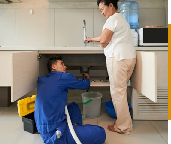 A plumber in blue overalls repairs a sink while a woman watches and runs the faucet.