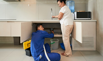 Plumber fixing a kitchen sink while a woman watches.